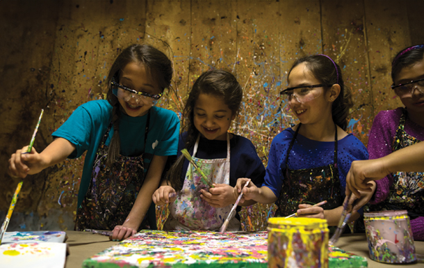Four girls dressed in aprons and safety goggles, in a paint-spattered room, painting